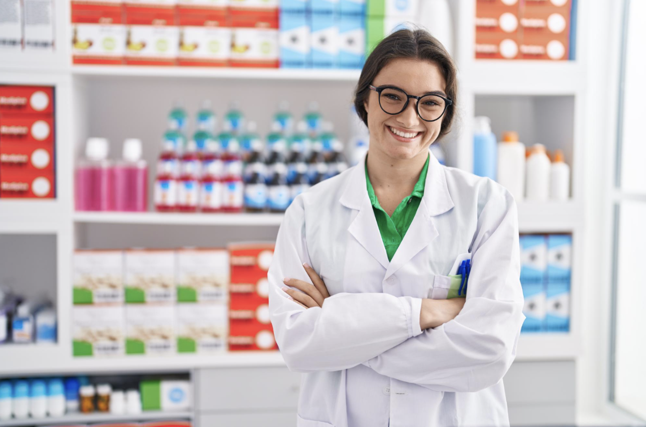 Smiling pharmacist in a white coat stands with arms crossed in a pharmacy with shelves of products behind her.